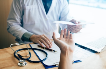 Foto de um médico, usando jaleco, entregando um papel, que é um atestado médico, para o paciente. Há um estetoscópio e uma caneta sobre a mesa de trabalho do médico. Não aparece o rosto dele nem o do paciente.