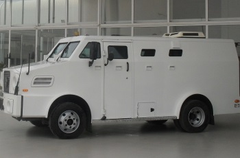 Foto de um carro-forte branco estacionado em frente à portaria de um prédio.