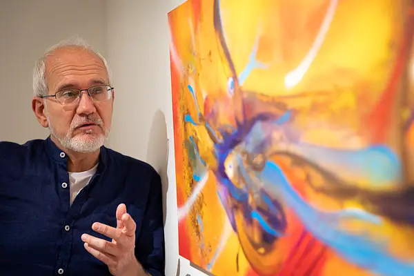 Foto do artista italiano Guido Boletti ao lado de uma de suas obras, gesticulando como se estivesse explicando o seu trabalho.