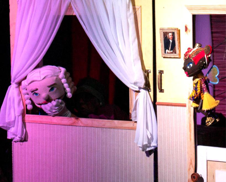 A imagem representa uma das cenas do espetáculo "O Menino Bach Visita o Brasil", e no centro da imagem esta o boneco de Johan Christian Bach apoiado em uma janela.