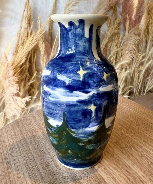 Vaso de cerâmica pintado à mão com paisagem noturna em azul, branco e verde. A decoração mostra estrelas, nuvens e pinheiros. O vaso está sobre uma mesa de madeira, com plantas secas desfocadas ao fundo