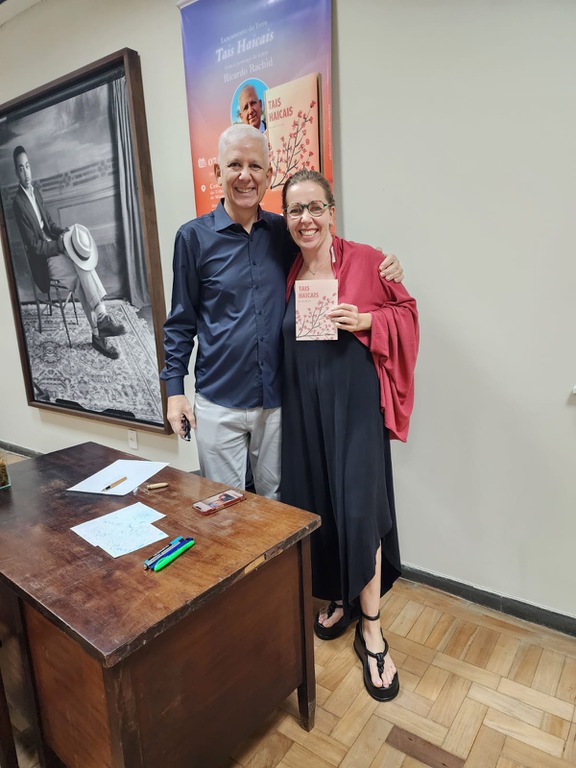ricardo rachid 2.jpg Fotografia do lançamento do livro Tais Haicais. O autor Ricardo Rachid, usando camisa azul de mangas compridas e calça clara, está sorrindo e abraçando uma participante do evento. Ela veste um vestido preto, um xale vermelho e sandálias, usa óculos, sorri para a câmera e segura uma cópia do livro Tais Haicais. À frente, há uma mesa de madeira com papéis, canetas e um celular. Ao fundo, vê-se um cartaz de divulgação do lançamento com a capa do livro e, à esquerda, um grande retrato em preto e branco de um homem sentado.