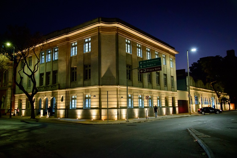 foto-centro-cultural.jpeg Fotografia noturna de um prédio histórico de esquina, iluminado por luzes amareladas. A fachada possui janelas retangulares e detalhes decorativos. Na calçada há placas de trânsito que indicam direções para a Avenida Amazonas, Avenida Pedro II e Praça Sete.