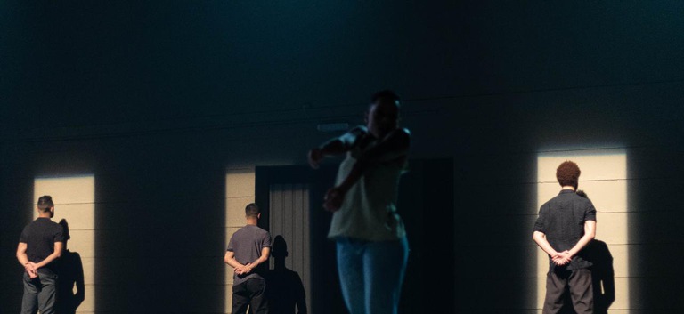 Uma cena do espetáculo com três homens virados de costas em frente a uma parede enquanto uma mulher dança ao centro em um ambiente pouco iluminado