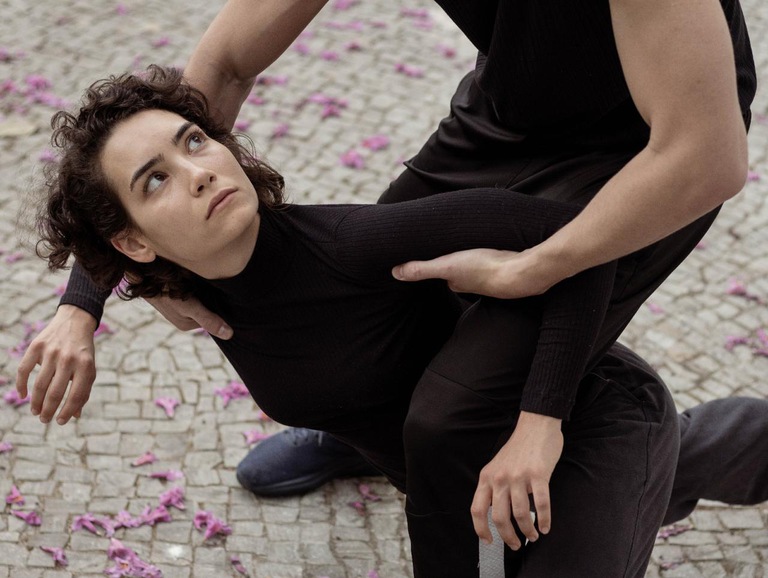 Cena do espetáculo em que há uma mulher vestida com uma roupa preta ajoelhada em um chão com pétalas de flores, sendo segurada por dois braços masculinos enquanto olha para cima