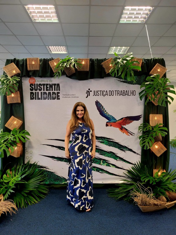 Júnia representando o TRT3 no evento, de vestido azul com branco, sem mangas. Atrás o folder do evento, branco, com uma arara vermelha.