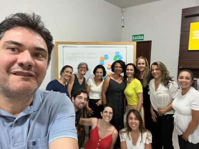 Participantes da oficina posam para a foto. São dez mulheres e um homem em torno de uma mesa de reunião, sorrindo, e cartazes colados nas paredes com post-its coloridos. 