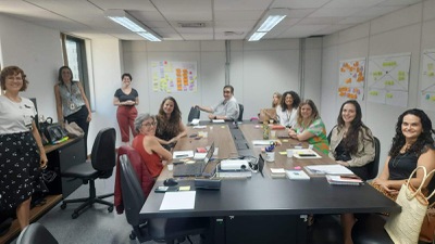 Um grupo de dez mulheres e um homem está em torno de uma mesa, a maioria sentados e três mulheres de pé, posando para a foto, enquanto nas paredes há canvas colados com post its. 