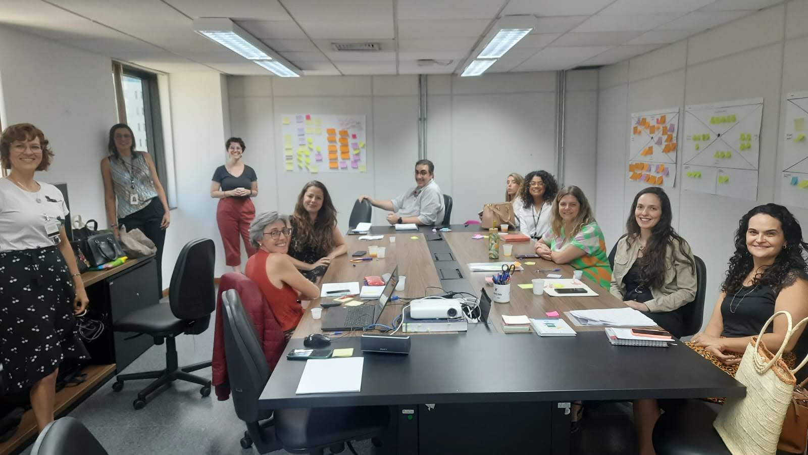 Participantes da oficina posam para a foto. São dez mulheres e um homem em torno de uma mesa de reunião, sorrindo, e cartazes colados nas paredes com post-its coloridos. 