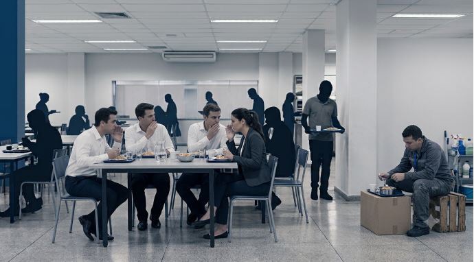 Imagem de um refeitório corporativo. Em primeiro plano, quatro pessoas de roupas sociais almoçam juntas e conversam à mesa. Ao fundo, outros trabalhadores circulam pelo ambiente. À direita, um funcionário uniformizado está sentado sozinho sobre uma caixa de papelão, fazendo sua refeição próximo aos materiais de limpeza, enquanto permanece afastado dos demais, evidenciando uma situação de isolamento e exclusão no ambiente de trabalho.