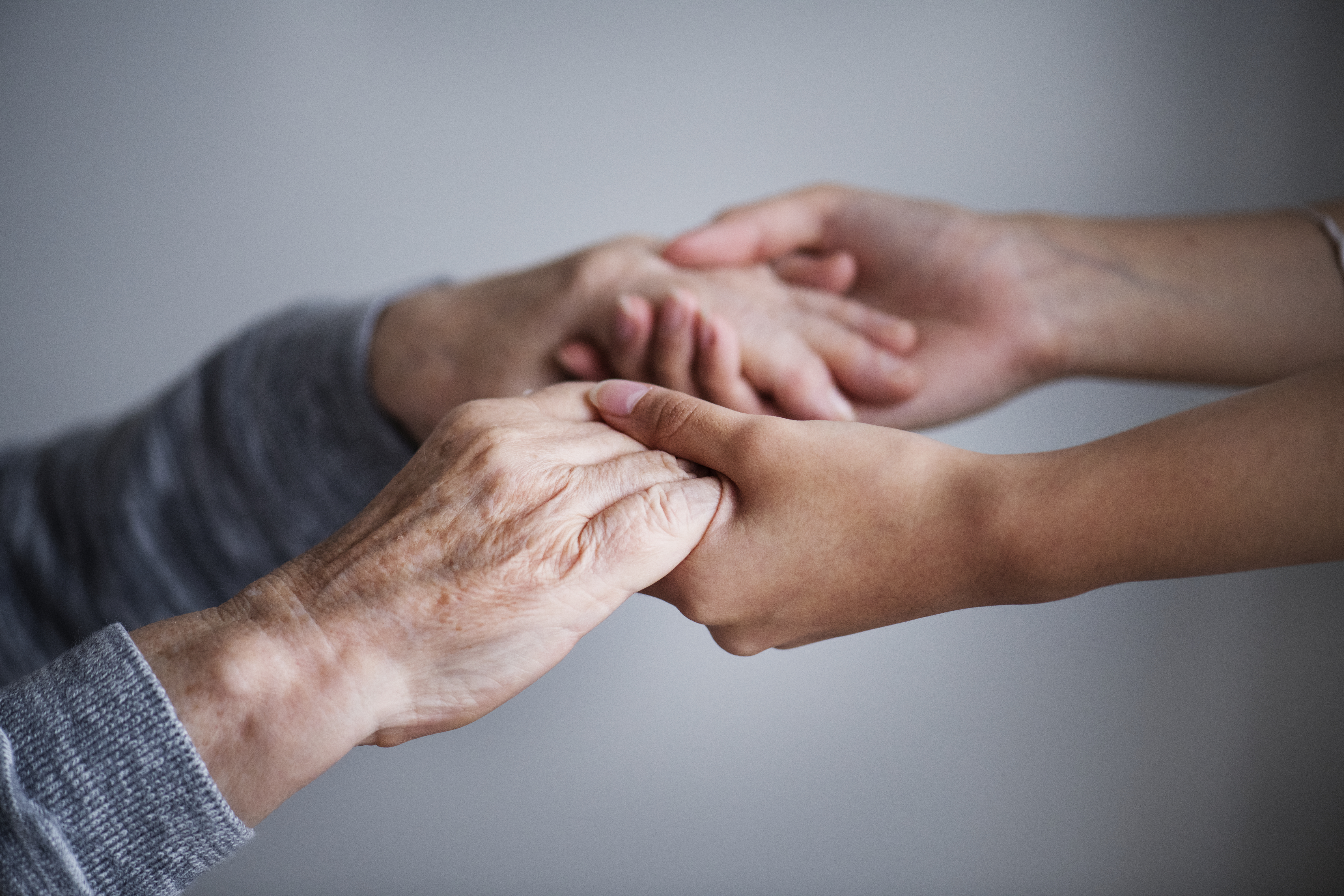 Close nas mãos de uma pessoa idosa sendo seguradas com cuidado por outra pessoa mais jovem. O gesto transmite acolhimento, respeito, empatia e valorização da experiência e da dignidade da pessoa idosa.