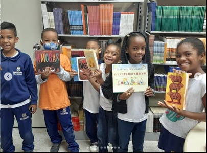 Foto de 6 crianças sorridentes, uniformizadas, em uma biblioteca, cada uma exibindo um livro doado