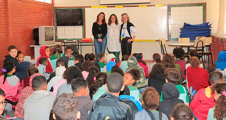Foto de em torno de 50 crianças pequenas assentadas no chão, em uma sala de aula, com a palestrante  e a juíza Cristiana Soares Campos na frente do quadro, de pé,  tendo ao lado esquerdo uma televisão. 