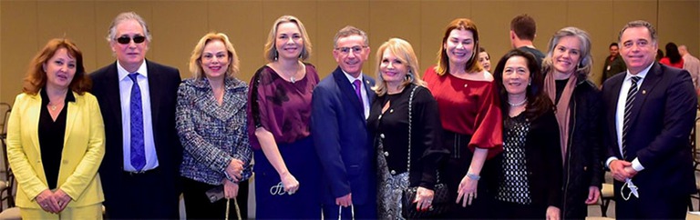 Foto de dez  participantes do evento de pé, sorridentes, à frente de cadeiras, em uma sala de piso acarpetado.